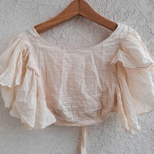 May include: A cream-colored, off-the-shoulder top with ruffled sleeves. The top is made of a lightweight fabric and has a relaxed fit.