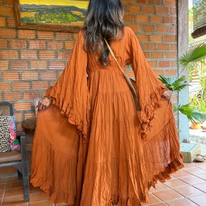 Op de afbeelding: Een roestkleurige maxi-jurk met lange, uitlopende mouwen en ruches. De jurk heeft een gelaagde rok en een getailleerde taille. Het model staat voor een bakstenen muur en draagt een crossbodytas.