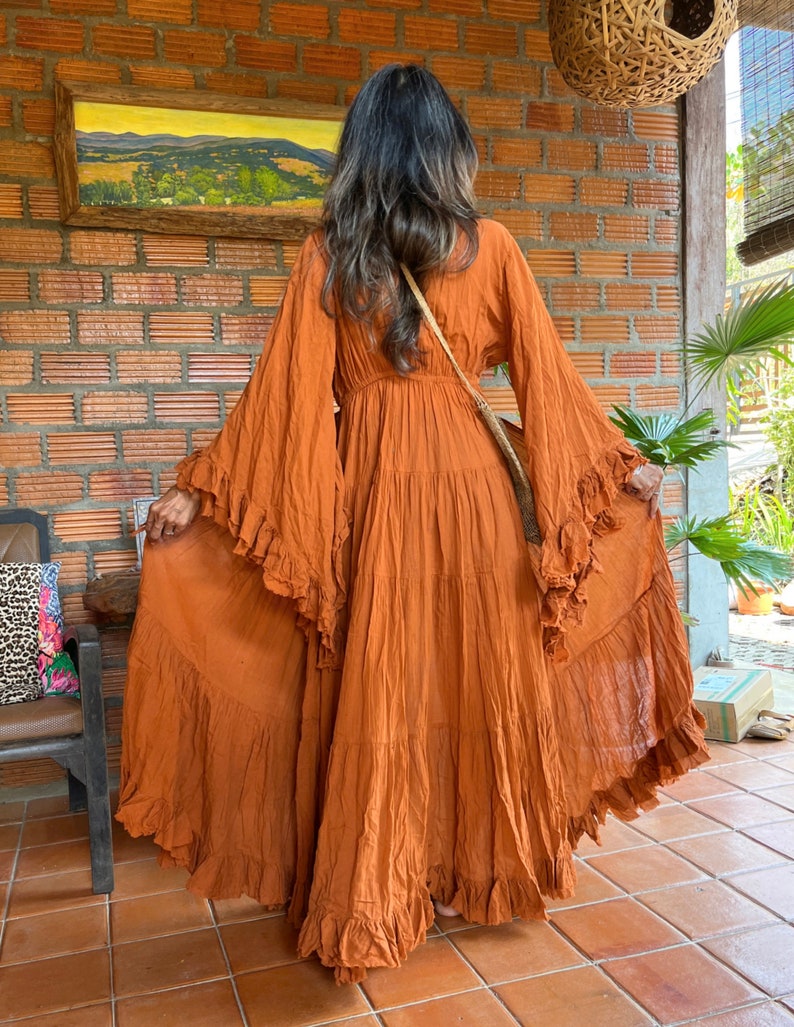 Op de afbeelding: Een roestkleurige maxi-jurk met lange, uitlopende mouwen en ruches. De jurk heeft een gelaagde rok en een getailleerde taille. Het model staat voor een bakstenen muur en draagt een crossbodytas.