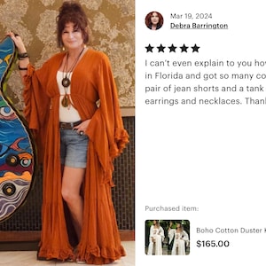 Op de afbeelding: Een vrouw draagt een roestkleurige duster kimono cardigan, staand naast een groot, kleurrijk gitaarvormig kunstwerk. De gitaar heeft een oogthema-ontwerp met blauwe, oranje en groene accenten. De cardigan is lang en vloeiend.