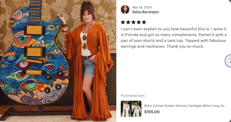 Op de afbeelding: Een vrouw draagt een roestkleurige duster kimono cardigan, staand naast een groot, kleurrijk gitaarvormig kunstwerk. De gitaar heeft een oogthema-ontwerp met blauwe, oranje en groene accenten. De cardigan is lang en vloeiend.