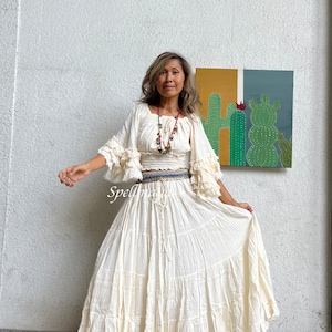 May include: A white, flowy maxi skirt with multiple tiers and a ruffled hem. The skirt is paired with a white, off-the-shoulder top with ruffled sleeves. The model is wearing a beaded necklace and a belt with multiple layers of colorful fabric.