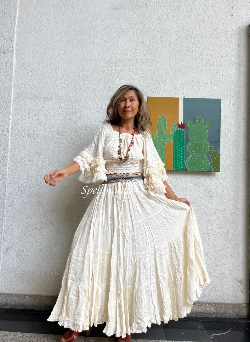 May include: A white, flowy maxi skirt with multiple tiers and a ruffled hem. The skirt is paired with a white, off-the-shoulder top with ruffled sleeves. The model is wearing a beaded necklace and a belt with multiple layers of colorful fabric.