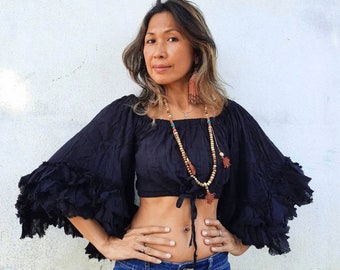 May include: A woman wearing a black off-the-shoulder crop top with ruffled sleeves and a brown beaded necklace. She is wearing blue jeans and has her hair down.