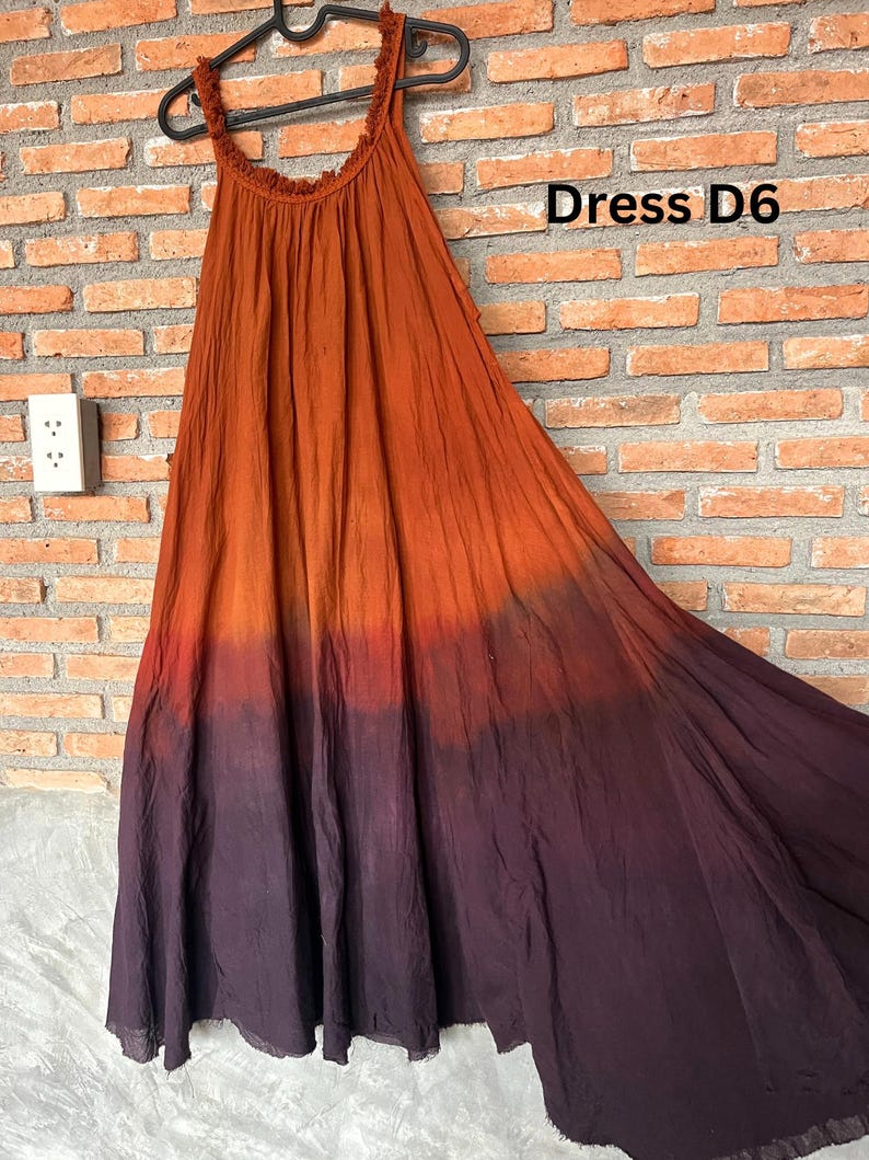 May include: A flowing, sleeveless dress with a gradient color scheme, transitioning from orange to deep purple. The dress is hanging on a black hanger against a brick wall. The text "Dress D6" is visible.