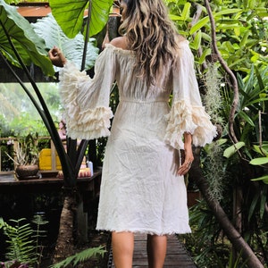 May include: A woman wearing a white off-the-shoulder dress with ruffled sleeves and a flowing skirt. She is walking on a wooden path in a lush green garden.