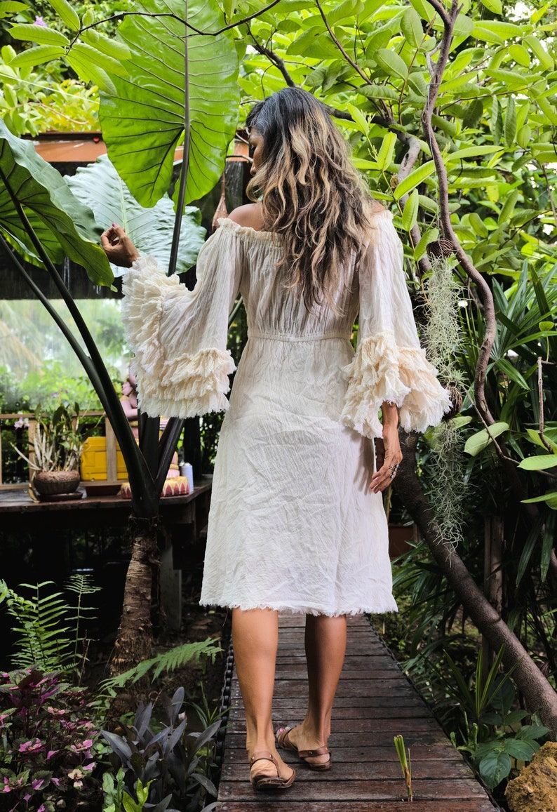 May include: A woman wearing a white off-the-shoulder dress with ruffled sleeves and a flowing skirt. She is walking on a wooden path in a lush green garden.