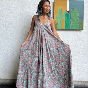 May include: A long, flowing maxi dress with a paisley print in shades of pink, blue, and brown. The dress has a V-neckline and thin straps. The model is wearing the dress with sandals.