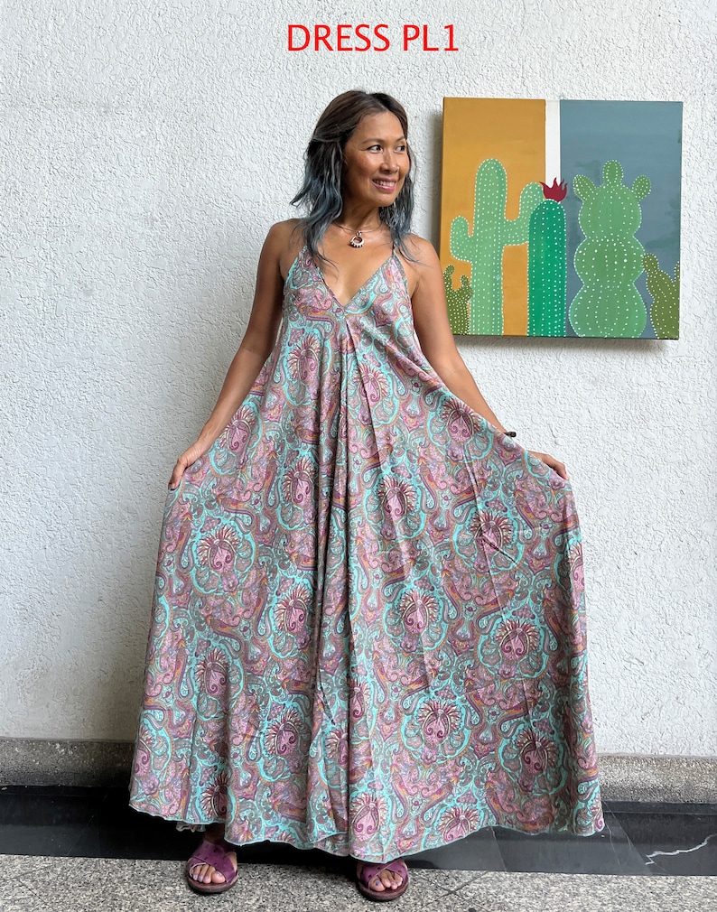 May include: A long, flowing maxi dress with a paisley print in shades of pink, blue, and brown. The dress has a V-neckline and thin straps. The model is wearing the dress with sandals.