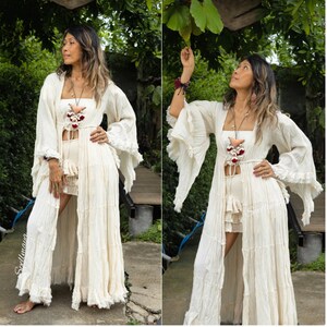 Op de afbeelding: Cr&egrave;mekleurige bohemien outfit met een lange, vloeiende duster met ruchesmouwen, een bijpassende top en shorts. De outfit is geaccessoireerd met een ketting en armbanden. De afbeelding is buiten gemaakt met weelderig groen op de achtergrond.