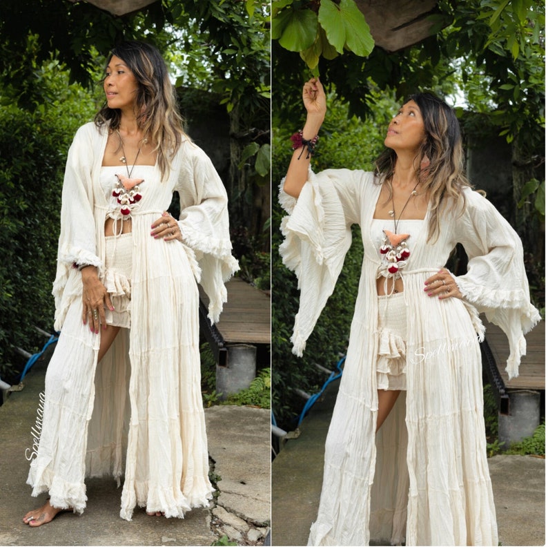 Op de afbeelding: Cr&egrave;mekleurige bohemien outfit met een lange, vloeiende duster met ruchesmouwen, een bijpassende top en shorts. De outfit is geaccessoireerd met een ketting en armbanden. De afbeelding is buiten gemaakt met weelderig groen op de achtergrond.