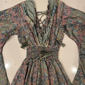 May include: A long-sleeved dress with a paisley print in shades of green, blue, and pink. The dress has a lace-up detail in the front and a cinched waist with a tie.