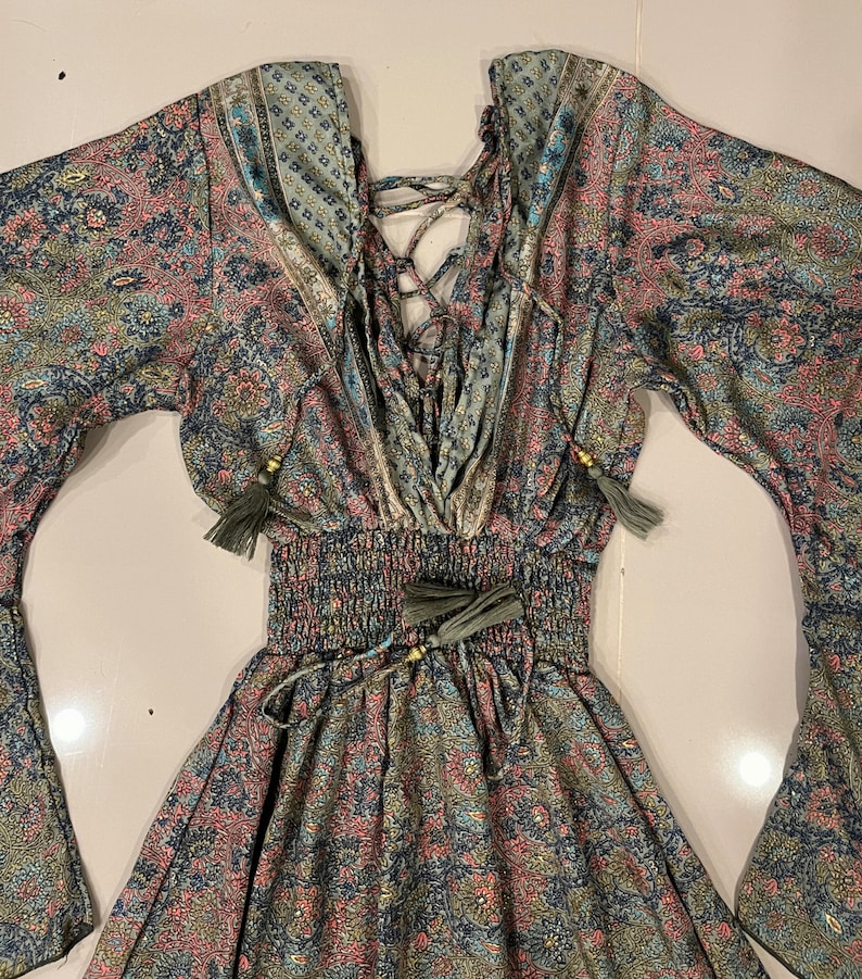 May include: A long-sleeved dress with a paisley print in shades of green, blue, and pink. The dress has a lace-up detail in the front and a cinched waist with a tie.