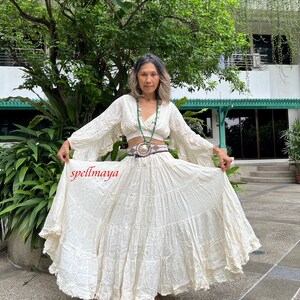 May include: A white, tiered maxi skirt with a ruffled hem. The skirt is made of a lightweight, flowy fabric and has a relaxed fit. The skirt is paired with a white, cropped top with long, bell sleeves. The top has a V-neckline and a button closure at the front. The model is wearing a green beaded necklace and a brown belt with a silver buckle. The text "spellmaya" is visible in the lower left corner of the image.