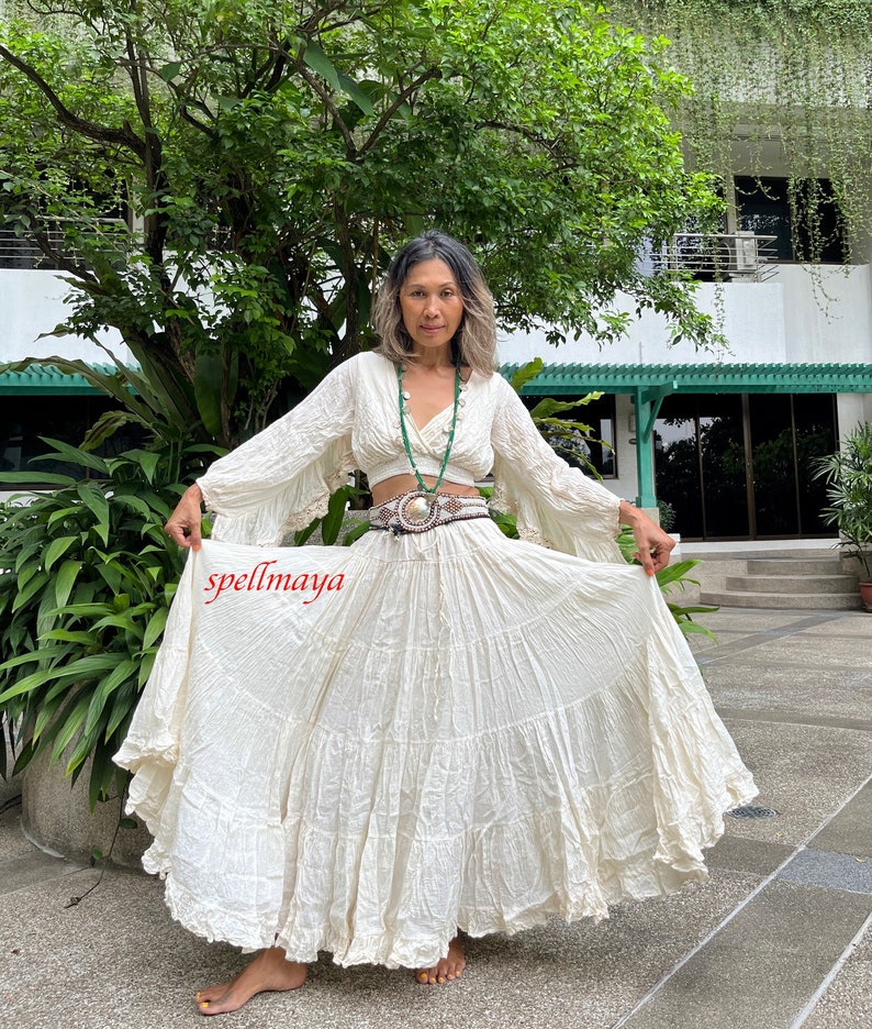 May include: A white, tiered maxi skirt with a ruffled hem. The skirt is made of a lightweight, flowy fabric and has a relaxed fit. The skirt is paired with a white, cropped top with long, bell sleeves. The top has a V-neckline and a button closure at the front. The model is wearing a green beaded necklace and a brown belt with a silver buckle. The text "spellmaya" is visible in the lower left corner of the image.