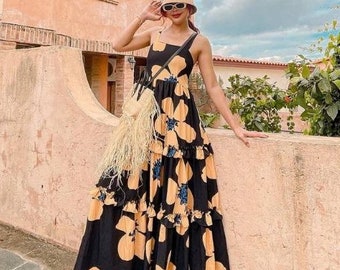 May include: A black maxi dress with a yellow floral pattern and ruffled tiers. The dress has a square neckline and thin straps. The person is wearing a straw hat, sunglasses, and sneakers. A straw bag is hanging from the shoulder.