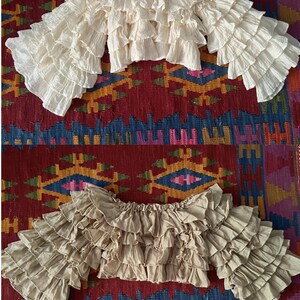 Pode incluir: Duas blusas de ombro a ombro com mangas com babados. Uma &eacute; creme, a outra &eacute; bege. As blusas s&atilde;o feitas de um tecido leve e t&ecirc;m v&aacute;rias camadas de babados. O fundo &eacute; um tapete estampado colorido.