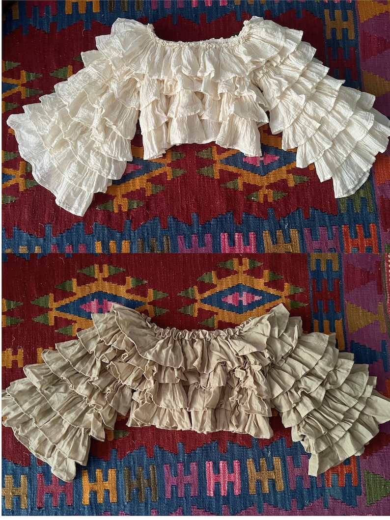 Pode incluir: Duas blusas de ombro a ombro com mangas com babados. Uma &eacute; creme, a outra &eacute; bege. As blusas s&atilde;o feitas de um tecido leve e t&ecirc;m v&aacute;rias camadas de babados. O fundo &eacute; um tapete estampado colorido.