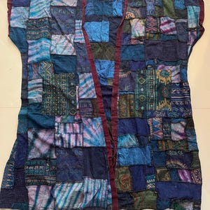 May include: A sleeveless open-front vest featuring a patchwork design in various shades of blue, teal, and purple. The garment displays diverse patterns, including geometric shapes, tie-dye, and script text. It has a dark maroon trim. Style BU4.