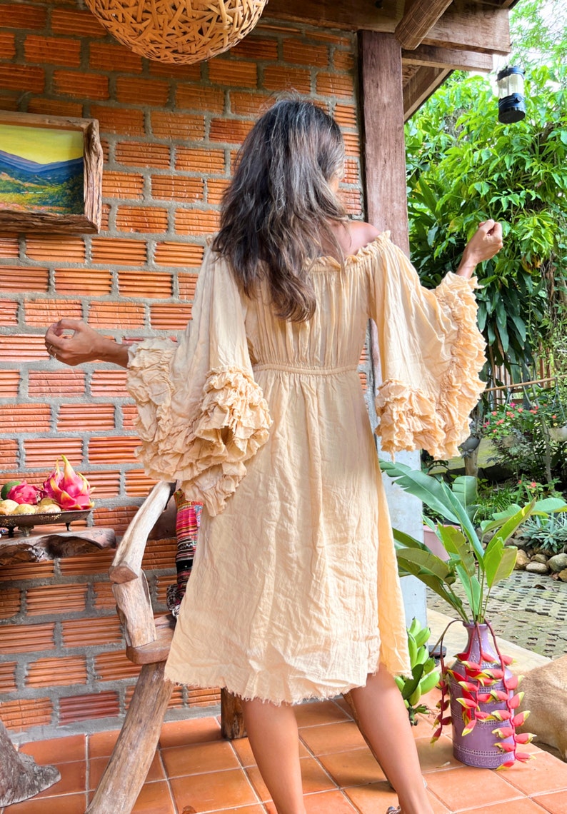 May include: A light beige off-the-shoulder dress with ruffled sleeves and a gathered waist. The dress is made of a lightweight, flowy fabric and has a relaxed fit.