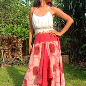 May include: A woman wearing a white crochet top and a long, flowing red and pink skirt with a floral print. The skirt has a high-low hemline and is tied at the waist.
