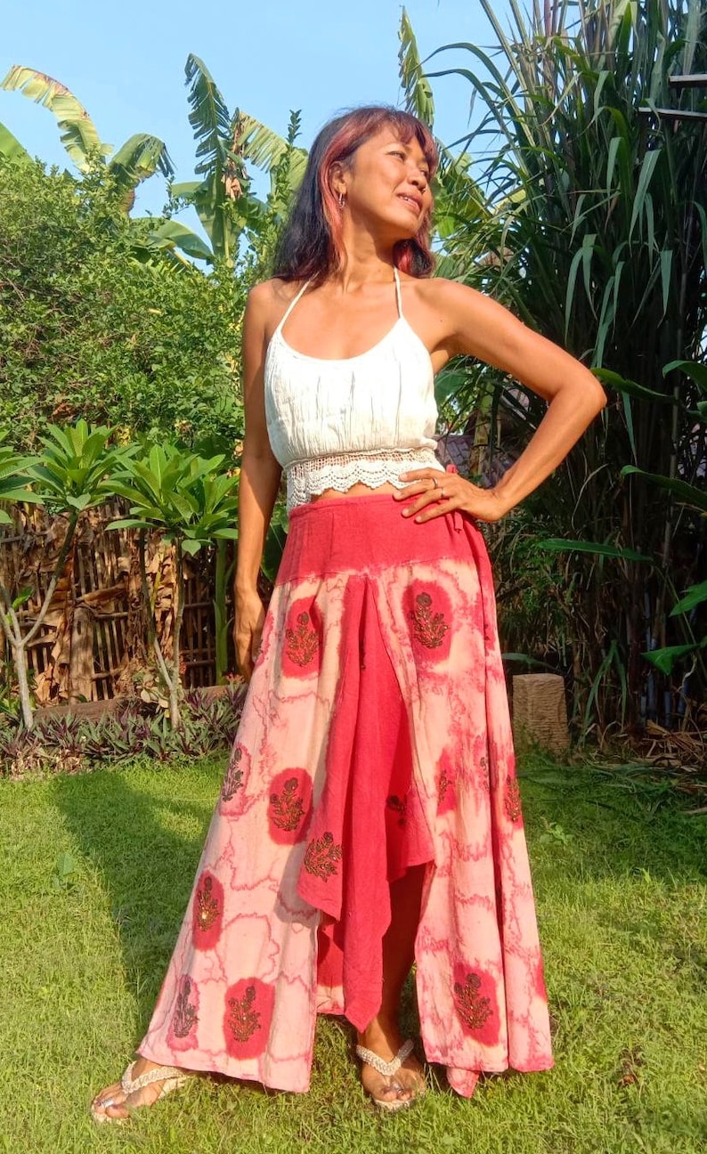 May include: A woman wearing a white crochet top and a long, flowing red and pink skirt with a floral print. The skirt has a high-low hemline and is tied at the waist.