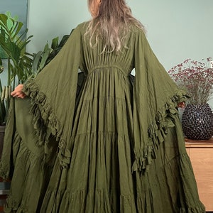 May include: Olive green maxi dress with tiered skirt and ruffled sleeves. The dress features a cinched waist and flowing fabric, creating a bohemian style. The model is wearing the dress in a room with plants and a wooden cabinet.