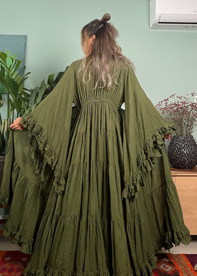 May include: Olive green maxi dress with tiered skirt and ruffled sleeves. The dress features a cinched waist and flowing fabric, creating a bohemian style. The model is wearing the dress in a room with plants and a wooden cabinet.