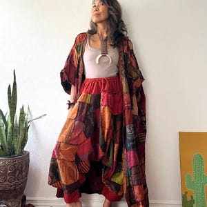 May include: A light beige tank top, a long patchwork kimono, and loose-fitting patchwork trousers. The garments feature a variety of warm-toned fabric squares in shades of red, orange, and brown, with various printed patterns and symbols throughout.