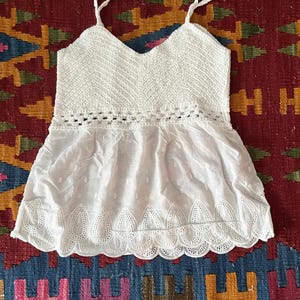 May include: White camisole top featuring a crochet bodice and a flowing, eyelet-detailed bottom. The top has spaghetti straps and a V-neck. The word "White" is printed in white text at the top of the image. The top is displayed on a patterned rug.