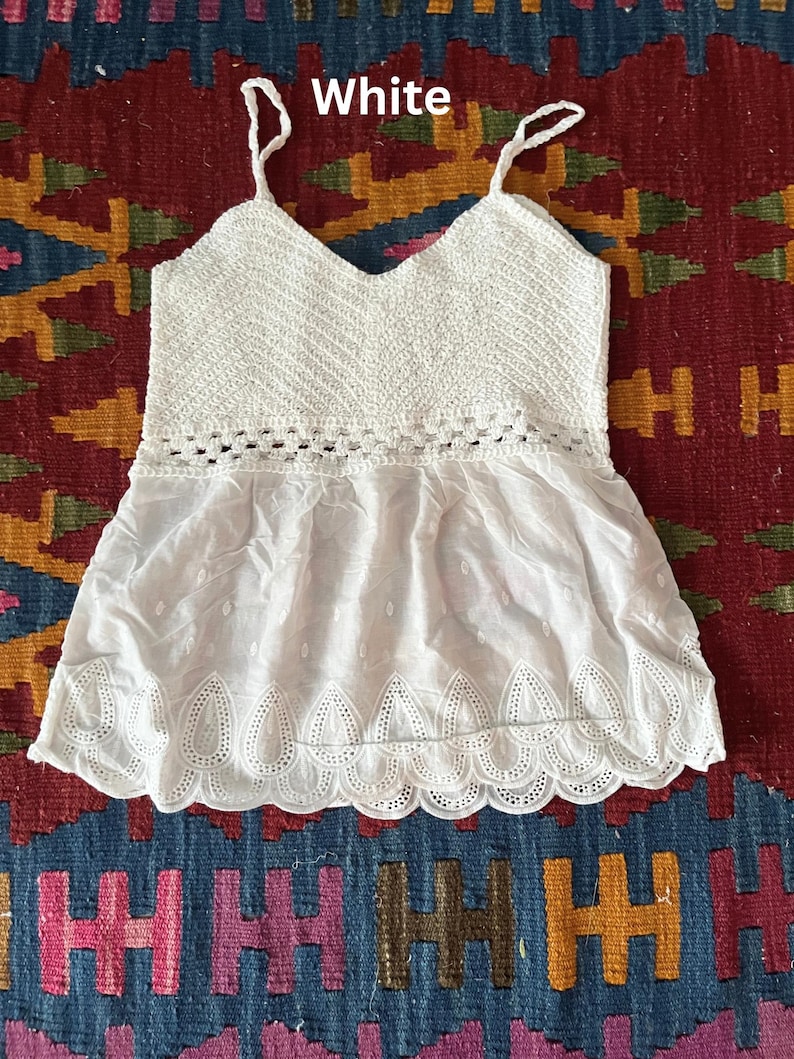 May include: White camisole top featuring a crochet bodice and a flowing, eyelet-detailed bottom. The top has spaghetti straps and a V-neck. The word "White" is printed in white text at the top of the image. The top is displayed on a patterned rug.