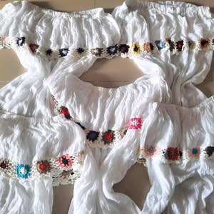 May include: Four white off-the-shoulder tops with colorful crocheted trim. The tops are made of a lightweight fabric and have a relaxed fit.