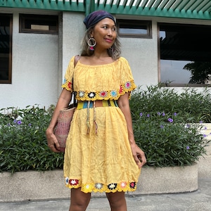 May include: A yellow crochet dress with colorful trim and a matching belt. The dress has a square neckline and short sleeves. The model is wearing a blue bandana and silver hoop earrings.