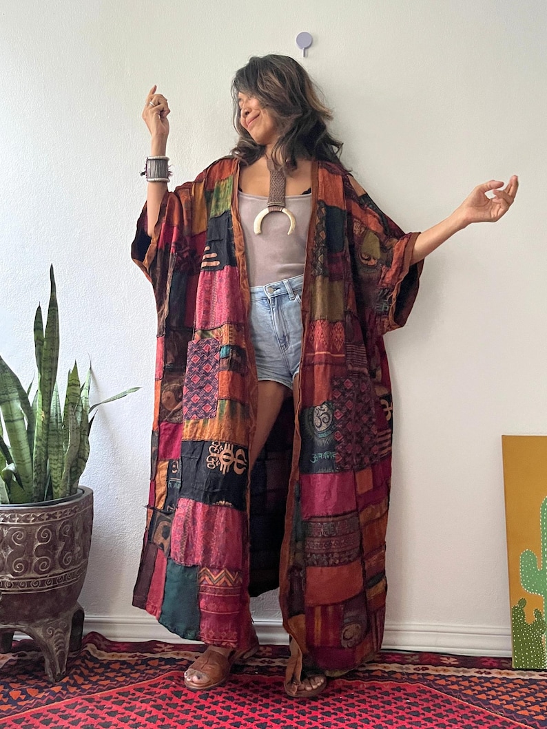 May include: A long, open-front kimono featuring a patchwork design with various fabric squares in shades of red, orange, brown, black, and green. The garment displays diverse patterns, including geometric shapes and symbols, with wide sleeves and a loose, flowing fit.