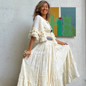 May include: A white, tiered maxi skirt with a ruffled hem. The skirt is made of a lightweight, flowy fabric and has a relaxed fit. The model is wearing the skirt with a white, ruffled top and a beaded belt.