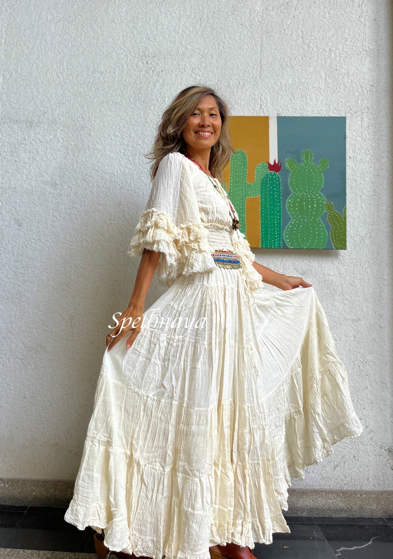 May include: A white, tiered maxi skirt with a ruffled hem. The skirt is made of a lightweight, flowy fabric and has a relaxed fit. The model is wearing the skirt with a white, ruffled top and a beaded belt.