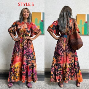 May include: Two women wearing long, flowing maxi dresses with a tie-dye pattern in shades of orange, red, black, and yellow. The dresses have a ruffled hemline and are cinched at the waist with a belt. The women are standing in front of a white wall with colorful artwork behind them. The text "STYLES" is written in red letters at the top of the image.