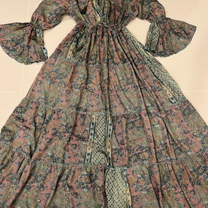 May include: A long-sleeved maxi dress with a V-neckline and a tie at the neck. The dress is made of a lightweight fabric with a paisley print in shades of blue, green, and pink. The dress has a tiered skirt and bell sleeves.