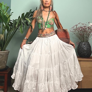 May include: A white tiered maxi skirt with a ruffled hem. The skirt is worn with a green and brown patterned crop top, a silver necklace, and a brown leather bag.