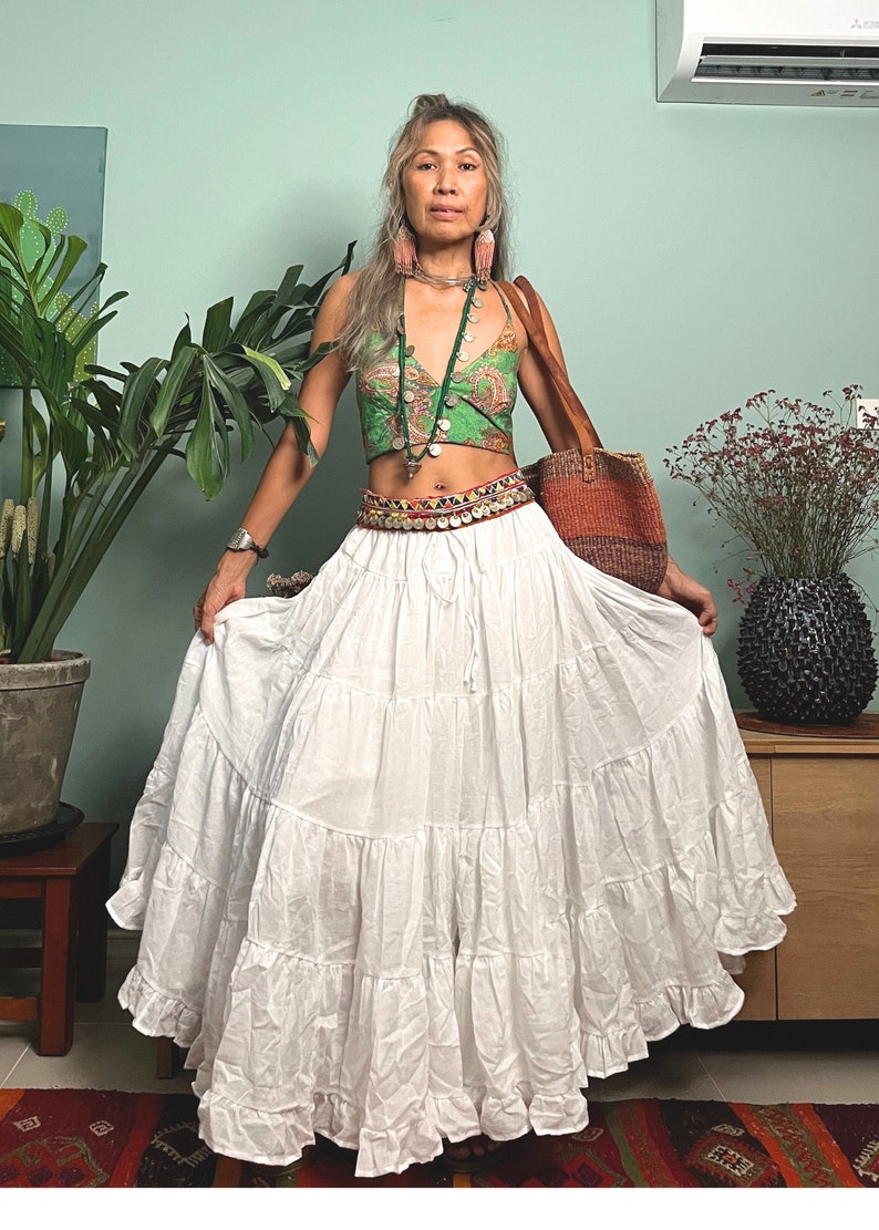 May include: A white tiered maxi skirt with a ruffled hem. The skirt is worn with a green and brown patterned crop top, a silver necklace, and a brown leather bag.