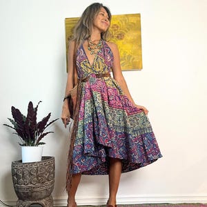 May include: A woman wearing a colorful patterned halter dress with a brown leather belt. She is standing on a patterned rug and has a brown leather bag slung over her shoulder.
