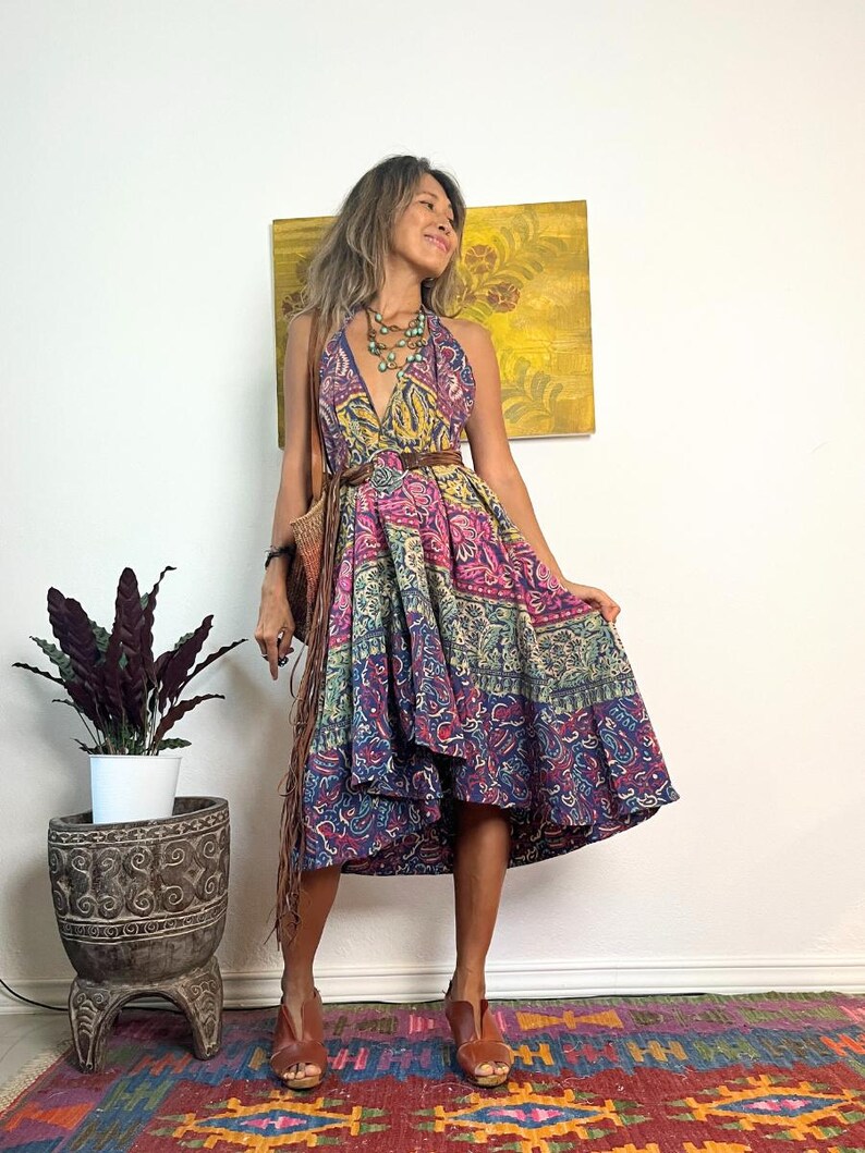 May include: A woman wearing a colorful patterned halter dress with a brown leather belt. She is standing on a patterned rug and has a brown leather bag slung over her shoulder.