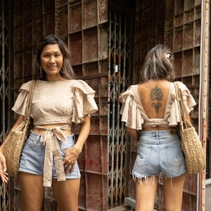 May include: A woman wearing a light brown linen top with a tie at the waist and light blue denim shorts. The top has a ruffled design on the sleeves and back. The woman is standing in front of a metal gate.