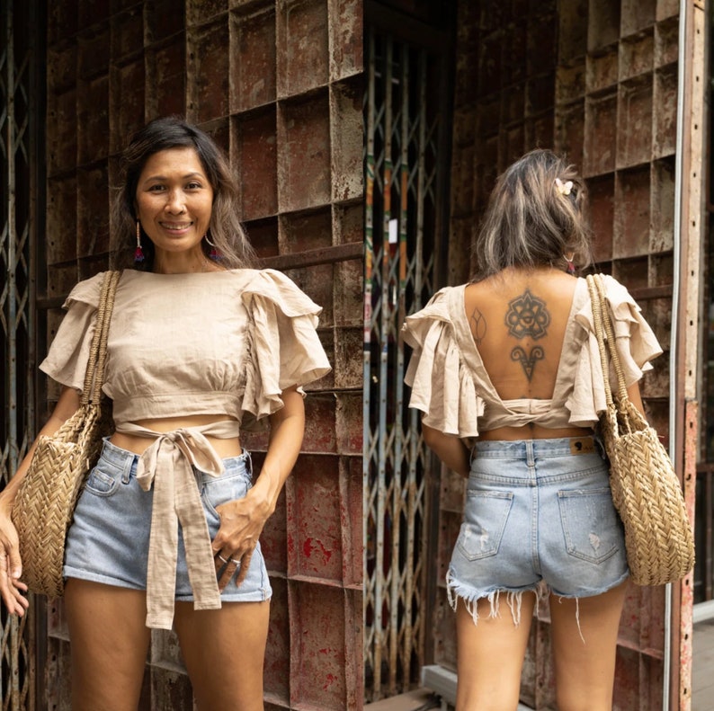May include: A woman wearing a light brown linen top with a tie at the waist and light blue denim shorts. The top has a ruffled design on the sleeves and back. The woman is standing in front of a metal gate.