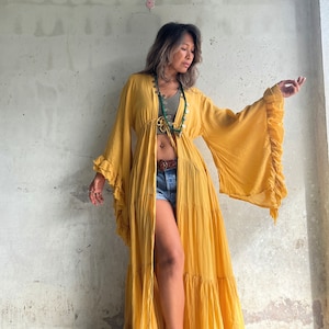 May include: Mustard yellow maxi dress with long, flared sleeves and ruffled edges. The dress features a cinched waist and a flowing skirt. A green beaded necklace and a brown belt are also visible.
