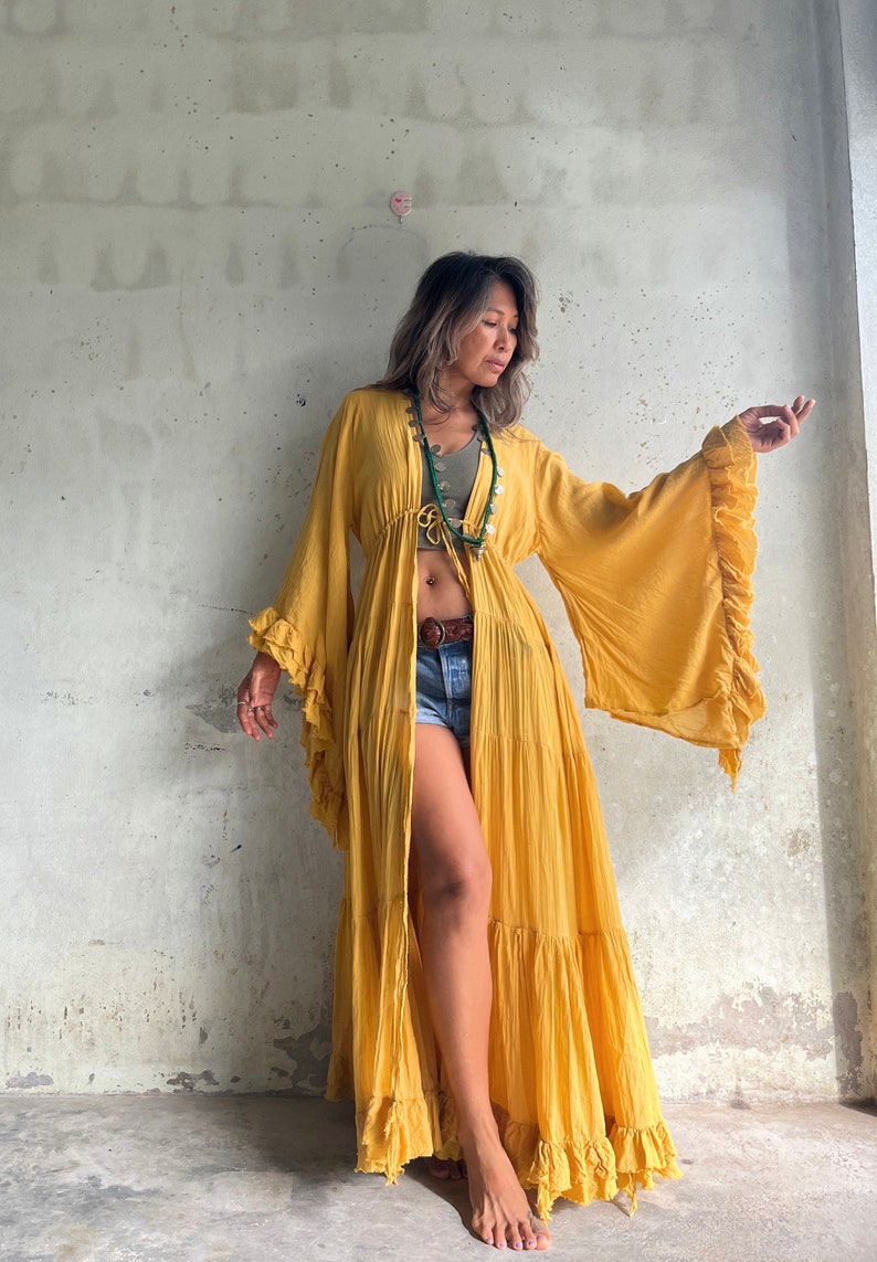 May include: Mustard yellow maxi dress with long, flared sleeves and ruffled edges. The dress features a cinched waist and a flowing skirt. A green beaded necklace and a brown belt are also visible.