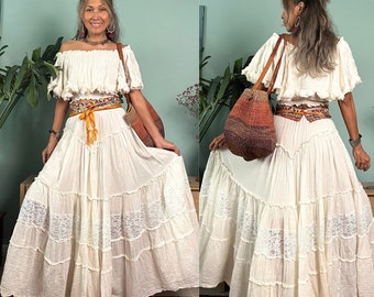 May include: A white off-the-shoulder peasant top with a ruffled neckline and a white tiered maxi skirt with lace trim. The skirt has a brown leather belt with a colorful woven design. The model is holding a woven bag with a brown leather strap.