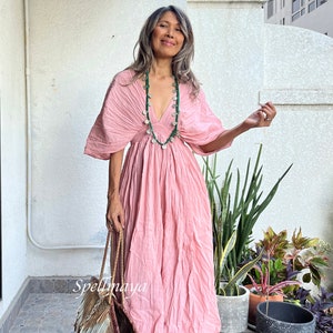 May include: A woman wearing a pink, flowing dress with a green beaded necklace. She is holding a woven bag with fringe and is standing in front of a white wall.