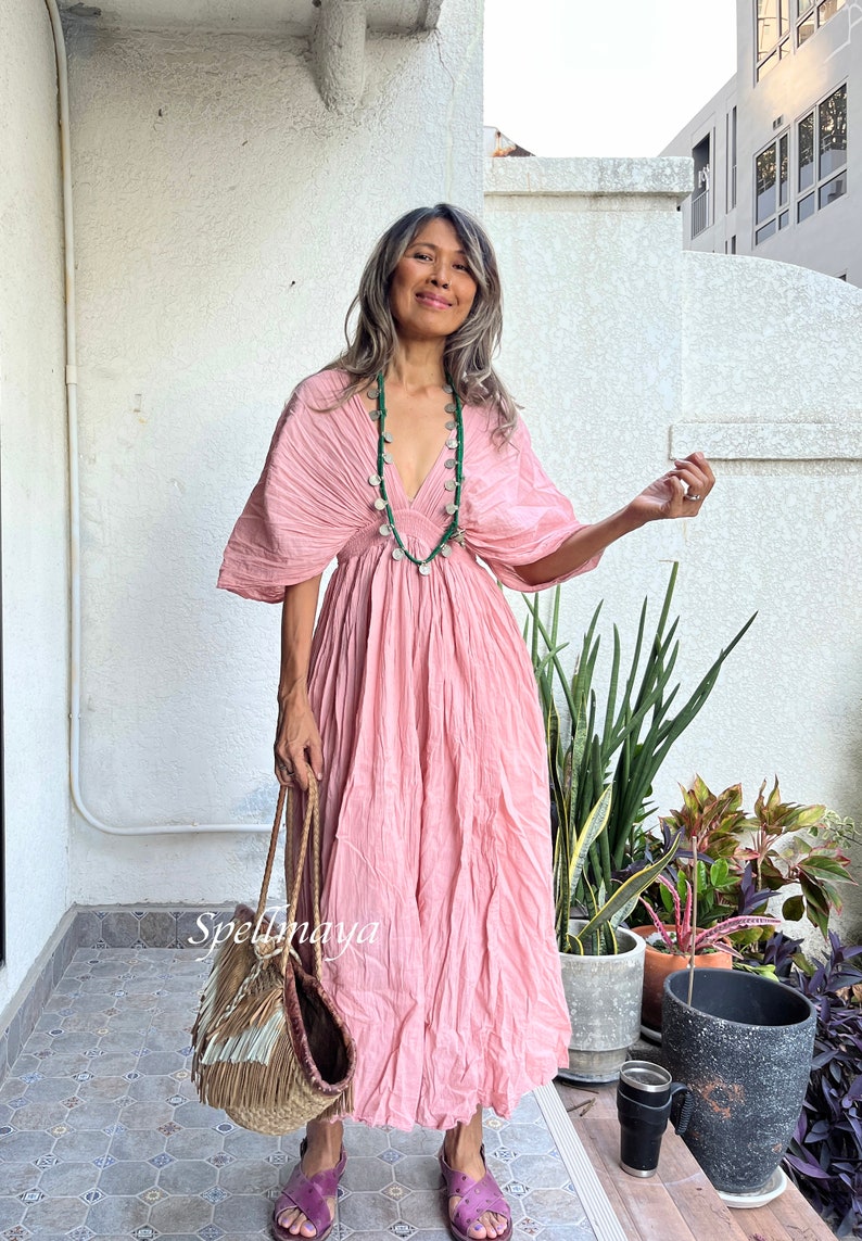 May include: A woman wearing a pink, flowing dress with a green beaded necklace. She is holding a woven bag with fringe and is standing in front of a white wall.