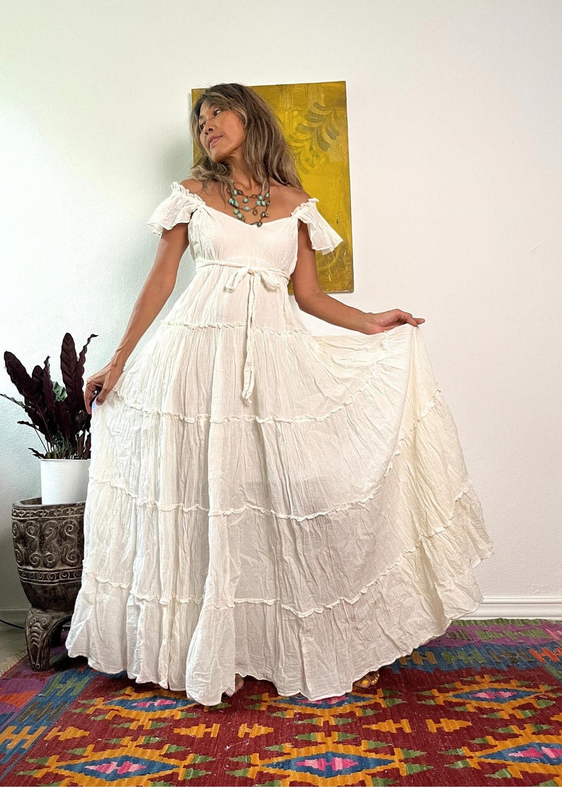 Pu&ograve; includere: Un abito maxi bianco a strati con un corpetto aderente e un laccio in vita. L'abito ha maniche corte a balze e una gonna fluida.